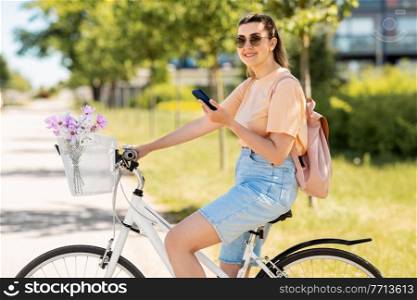 people, leisure and lifestyle - happy young woman with smartphone, backpack and flowers in basket of bicycle on city street. woman with smartphone on bicycle in city
