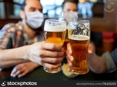 people, leisure and pandemic concept - male friends wearing face protective medical masks for protection from virus disease drinking beer and clinking glasses at bar or pub. male friends in masks drinking beer at bar or pub