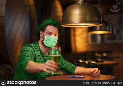 people, leisure and st patrick’s day concept - young man wearing face protective medical mask for protection from virus disease drinking green beer at bar or pub. young man in mask drinking green beer at bar