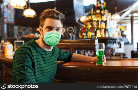 people, leisure and st patrick’s day concept - young man wearing face protective medical mask for protection from virus disease drinking green beer at bar or pub. young man in mask drinking green beer at bar
