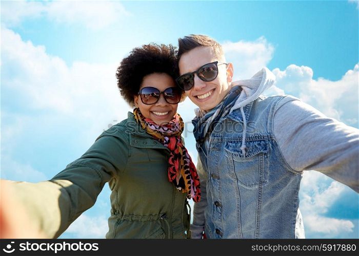 people, leisure and technology concept - happy international teenage couple taking selfie over blue sky and clouds background