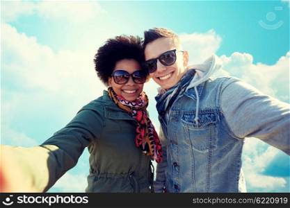 people, leisure and technology concept - happy international teenage couple taking selfie over blue sky and clouds background