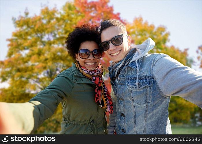 people, leisure and technology concept - happy mixed race teenage international couple taking selfie over autumn park background. happy mixed race couple taking selfie in autumn