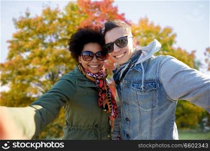 people, leisure and technology concept - happy mixed race teenage international couple taking selfie over autumn park background. happy mixed race couple taking selfie in autumn