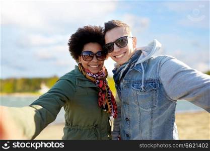people, leisure and technology concept - happy mixed-race teenage taking selfie over beach background. happy teenage couple taking selfie over beach