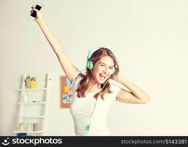 people, leisure and technology concept - happy woman or teenage girl in headphones listening to music from smartphone and dancing on bed at home. happy woman in headphones ihaving fun at home