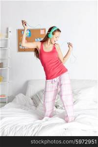 people, leisure and technology concept - happy woman or teenage girl in headphones listening to music from smartphone and dancing on bed at home