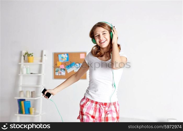 people, leisure and technology concept - happy woman or teenage girl in headphones listening to music from smartphone and dancing on bed at home