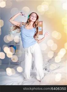people, leisure and technology concept - happy woman or teenage girl in headphones listening to music from smartphone and dancing on bed at home. happy woman in headphones ihaving fun at home