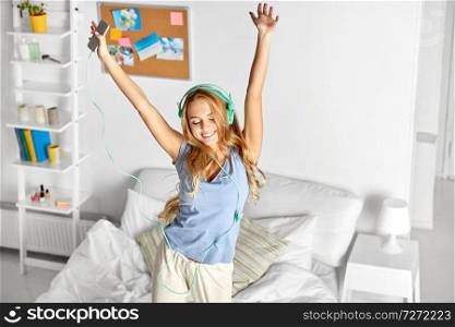 people, leisure and technology concept - happy woman or teenage girl in headphones listening to music from smartphone and dancing on bed at home. happy woman in headphones having fun at home