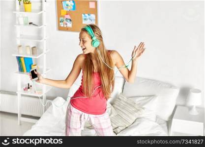 people, leisure and technology concept - happy woman or teenage girl in headphones listening to music from smartphone and dancing on bed at home. happy woman in headphones having fun at home