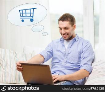 people, leisure and technology concept - smiling young man with laptop computer and trolley icon shopping online at home