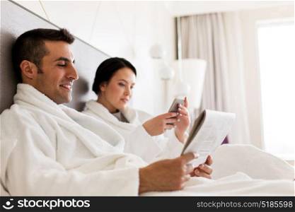 people, leisure and vacation concept - happy couple with newspaper and smartphone in bed at home or hotel room. happy couple in bed at home or hotel room