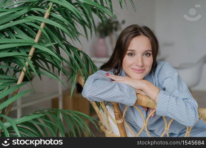 People, leisure concept. Beautiful European woman dressed in stylish clothes, sits on wooden chair near green plant, looks positively at camera, has day off, spends free time in family circle