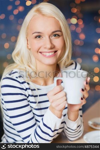 people, leisure, eating and drinking concept - happy young woman drinking tea or coffee at cafe or home over holidays lights background