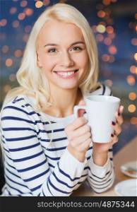 people, leisure, eating and drinking concept - happy young woman drinking tea or coffee at cafe or home over holidays lights background