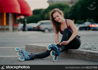 People leisure fitness sport recreation concept. Pleased young woman puts on rollerskates going to ride rolles in urban place has regular exercising goes in for dangerous sport adjustes laces