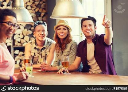 people, leisure, friendship and communication concept - group of happy smiling friends drinking beer and cocktails at bar or pub and pointing finger to something