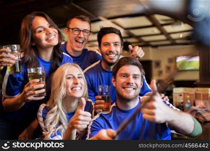 people, leisure, friendship and technology concept - happy friends or football fans taking picture by smartphone selfie stick, drinking beer and showing thumbs up at sport at bar or pub