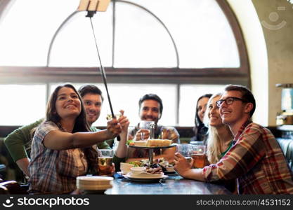 people, leisure, friendship and technology concept - happy friends taking picture by smartphone selfie stick, drinking beer and eating snacks at bar or pub