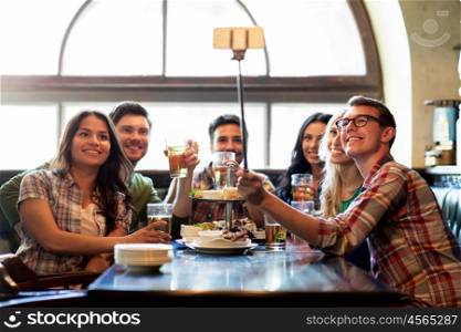 people, leisure, friendship and technology concept - happy friends taking picture by smartphone selfie stick, drinking beer and eating snacks at bar or pub