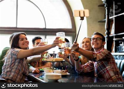people, leisure, friendship and technology concept - happy friends taking picture by smartphone selfie stick, drinking beer and eating snacks at bar or pub