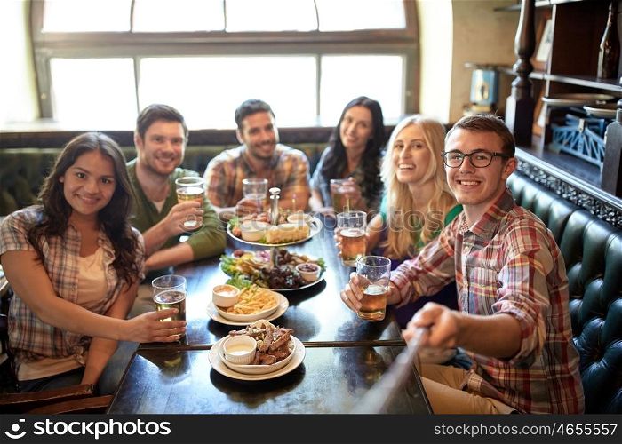 people, leisure, friendship and technology concept - happy friends taking picture by selfie stick, drinking beer and eating snacks at bar or pub