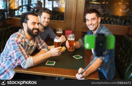 people, leisure, friendship, technology and bachelor party concept - happy male friends drinking beer and taking picture with smartphone selfie stick at bar or pub
