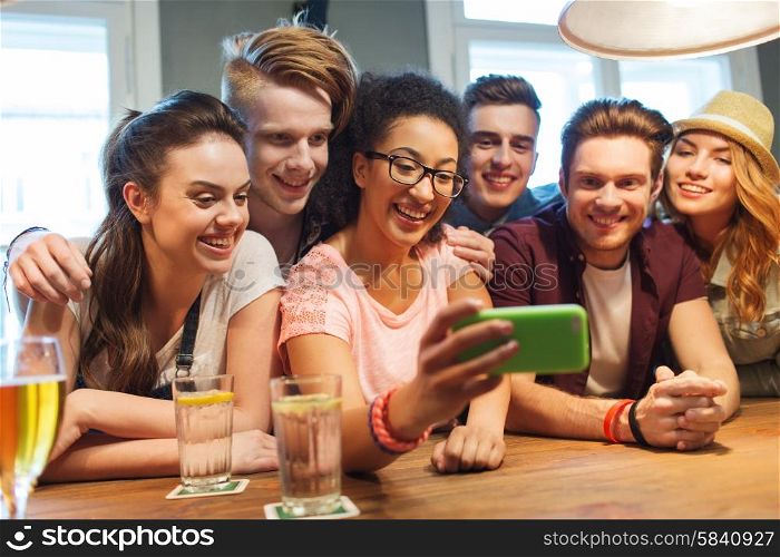 people, leisure, friendship, technology and communication concept - group of happy smiling friends with smartphone and drinks taking selfie at bar or pub