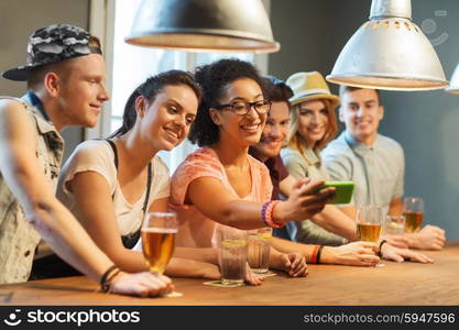 people, leisure, friendship, technology and communication concept - group of happy smiling friends with smartphone and drinks taking selfie at bar or pub