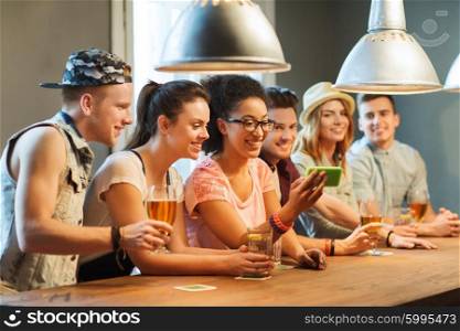 people, leisure, friendship, technology and communication concept - group of happy smiling friends with smartphone and drinks taking selfie at bar or pub