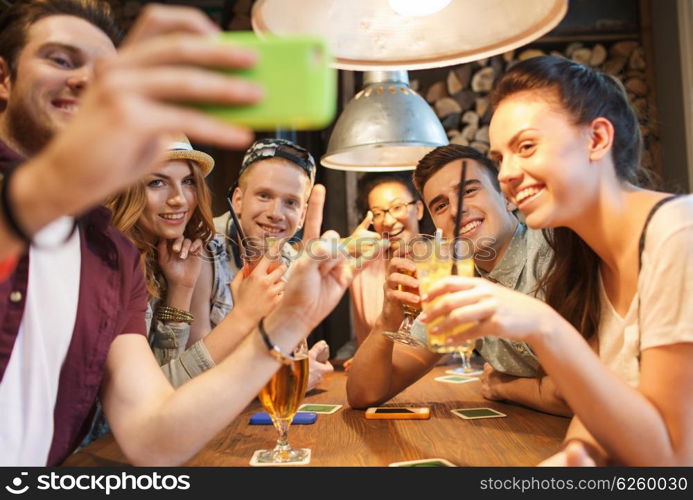 people, leisure, friendship, technology and communication concept - group of happy smiling friends with smartphone and drinks taking selfie at bar or pub