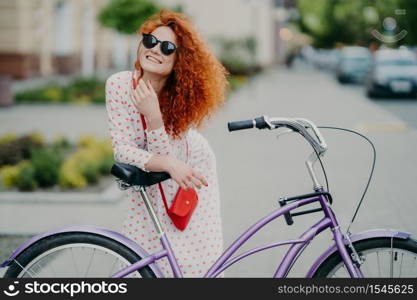 People, leisure, lifestyle and spare time concept. Cheerful curly woman focused into distance, rides bicycle in urban setting, enjoys her hobby, wears shades and long dress, has red small bag