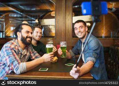 people, leisure, st patricks da and technology concept - happy male friends drinking beer and taking picture with smartphone selfie stick at bar or pub. friends taking selfie and drinking beer at bar