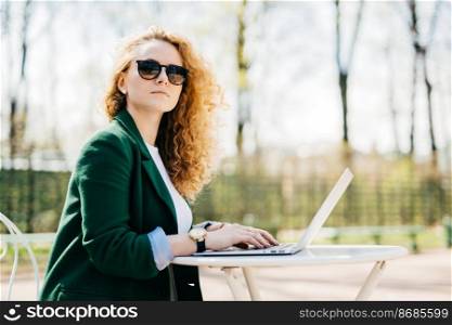 People, leisure, technology and communication. Beautiful businesswoman wearing sunglasses and elegant jacket using laptop computer to make conversation with her partners sitting sideways at white desk