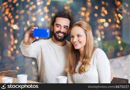 people, leisure, technology and dating concept - happy couple with smartphone taking selfie at cafe over holidays lights background