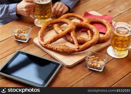 people, leisure, technology and drinks concept - close up of man drinking beer with pretzels, peanuts and tablet pc computer on table at bar or pub