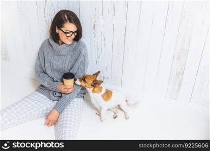 People, lifestyle and leisure concept. Carefree beautiful young woman dressed in domestic clothes, drinks takeaway coffee, spends free time with dog, enjoys calm atmosphere, being in good mood
