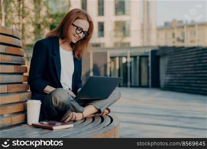 People lifestyle distance job and technology concept. Positive female blogger or freelancer types text of publication on laptop computer shares positive content online sits crossed legs on bench. Positive female blogger or freelancer types text of publication on laptop computer