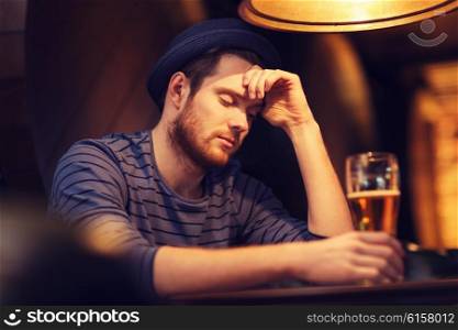 people, loneliness, alcohol and lifestyle concept - unhappy single young man in hat drinking beer at bar or pub