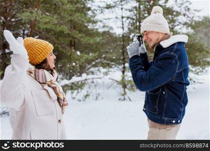 people, love and leisure concept - happy smiling man photographing woman in winter forest. happy man photographing woman in winter forest