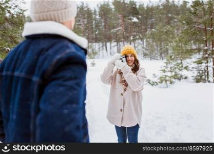 people, love and leisure concept - happy smiling woman photographing man in winter forest. happy woman photographing man in winter forest
