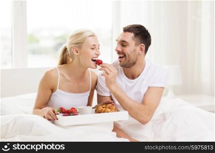 people, love, care and happiness concept - happy couple having breakfast in bed and eating strawberries at home