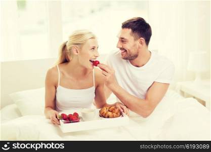 people, love, care and happiness concept - happy couple having breakfast in bed and eating strawberries at home