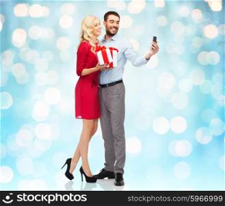people, love, couple, technology and holidays concept - happy young woman and man with birthday presents taking selfie by smartphone over blue lights background