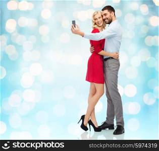 people, love, couple, technology and holidays concept - happy young woman and man taking selfie with smartphone and hugging over blue holidays lights background