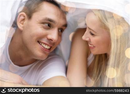people, love, relations and bedtime concept - happy couple under blanket in bed over lights background