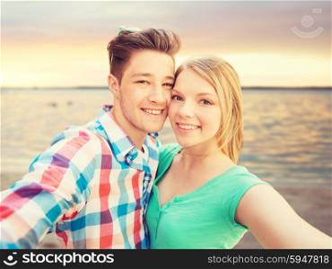 people, love, sea, technology and summer vacation concept - happy couple taking selfie with smartphone or camera over sunset beach background