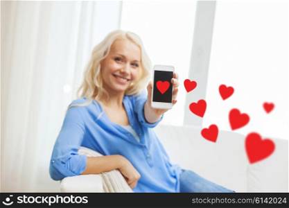 people, love, technology and communication concept - smiling woman showing smartphone with red hearts and sitting on couch at home
