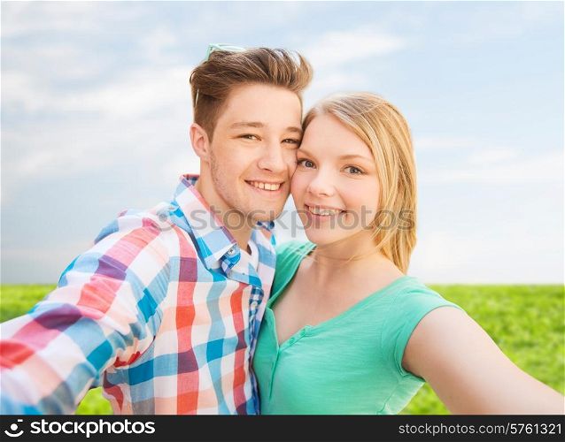 people, love, vacation, technology and summer concept - happy couple taking selfie with smartphone or camera over blue sky and grass background